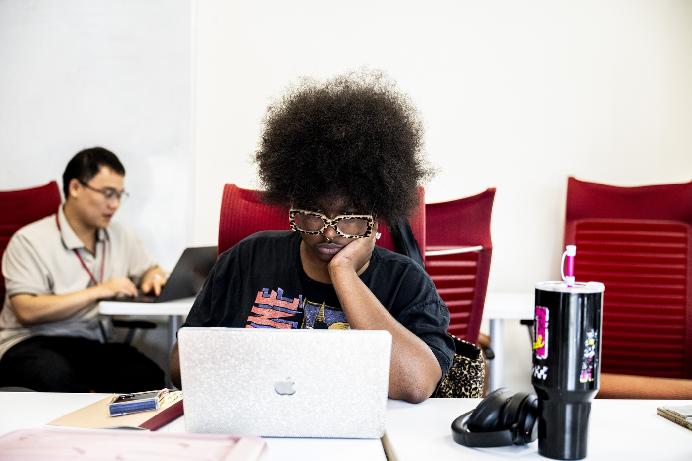 A student rests her chin on her hand while looking at her laptop, with another student working in the background.