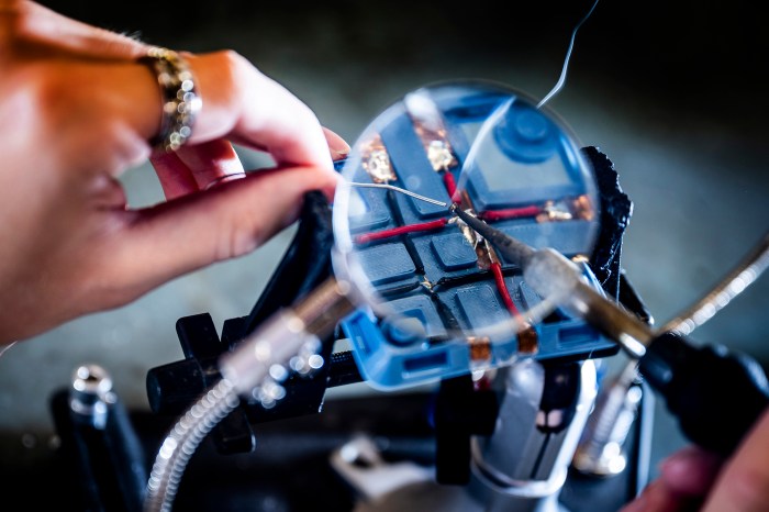A hand uses a heated tool to weld pieces together under a microscope.