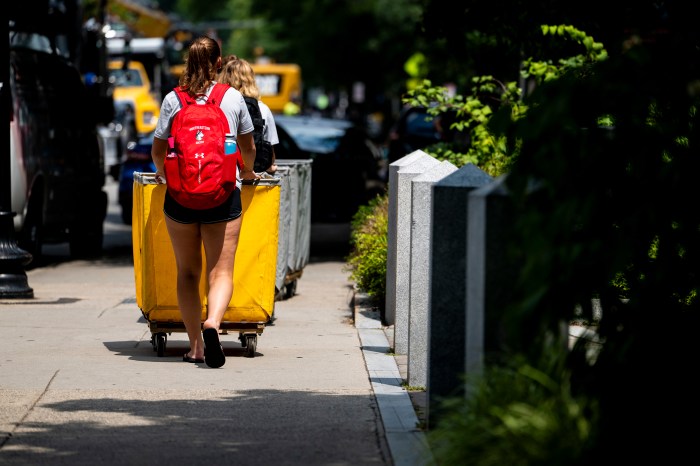Two Northeastern athletes, one wearing a red backpack, pull yellow move-in bins down a campus sidewalk.