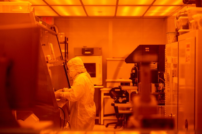 A researcher in a full cleanroom suit works among lab equipment under amber safety lighting in the Egan Research Center.