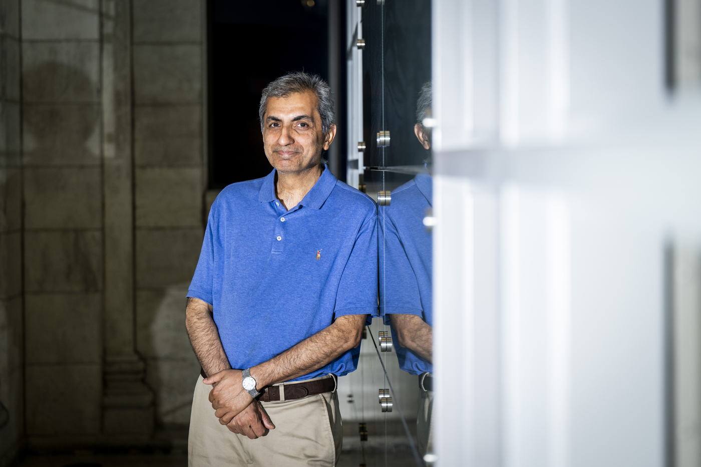 Hossein Mosallaei stands with hands clasped near a glass wall, his reflection visible beside him.