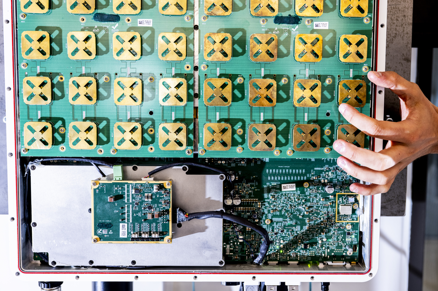 Close-up of a green circuit board with an array of gold-colored antenna modules, part of 5G and AI research equipment; a researcher's hand reaches toward it.