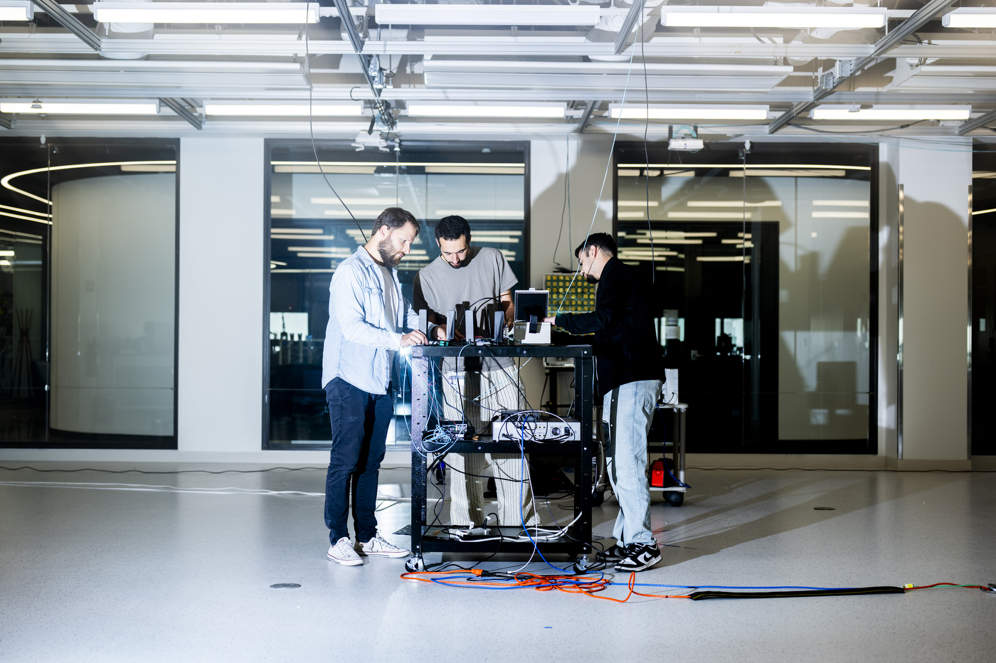 Three researchers — Michele Polese, Maxime Elkael, and Tamerlan Aghayev — collaborate around a wheeled equipment rack wired with cables in a bright lab space at Northeastern University's EXP building.