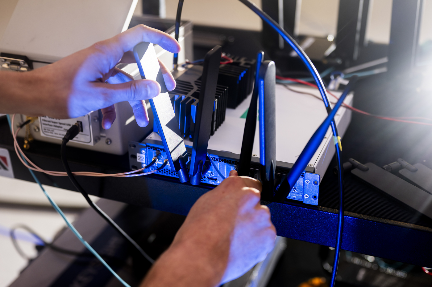 Close-up of a researcher's hands holding a small electronic device connected to blue-lit cables in a lab setup.