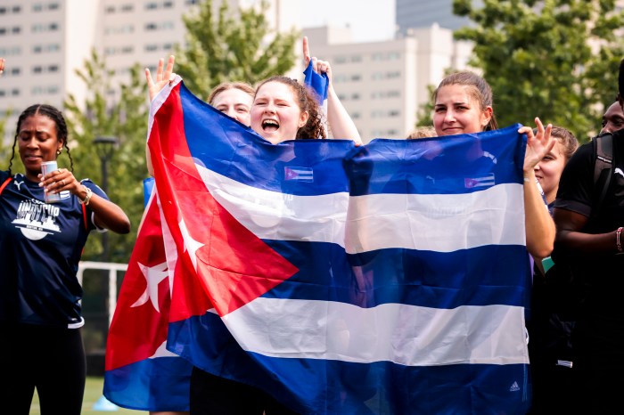A group of women celebrate and cheer while holding up a Cuban flag on a soccer field, with a smiling onlooker in the background.