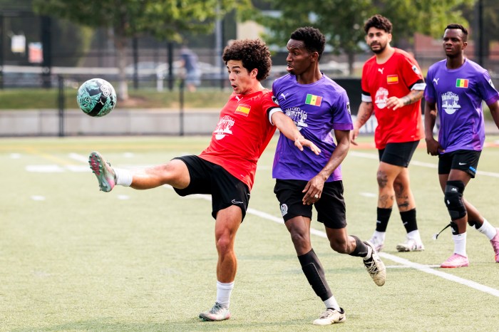 A soccer player in a red jersey kicks the ball while players in purple jerseys close in during a match.