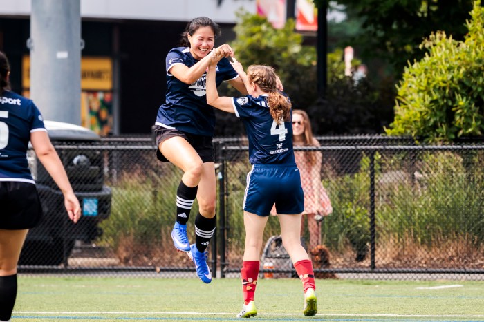 Two soccer players in navy jerseys celebrate together, jumping and clasping hands on the field.