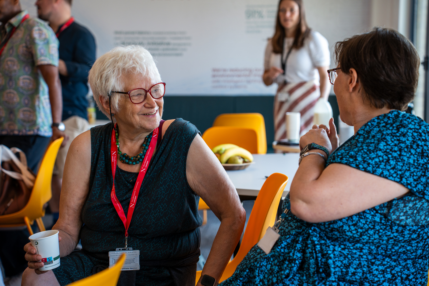 Two women talk while seated at a table, one wearing a lanyard, with others mingling in the background.