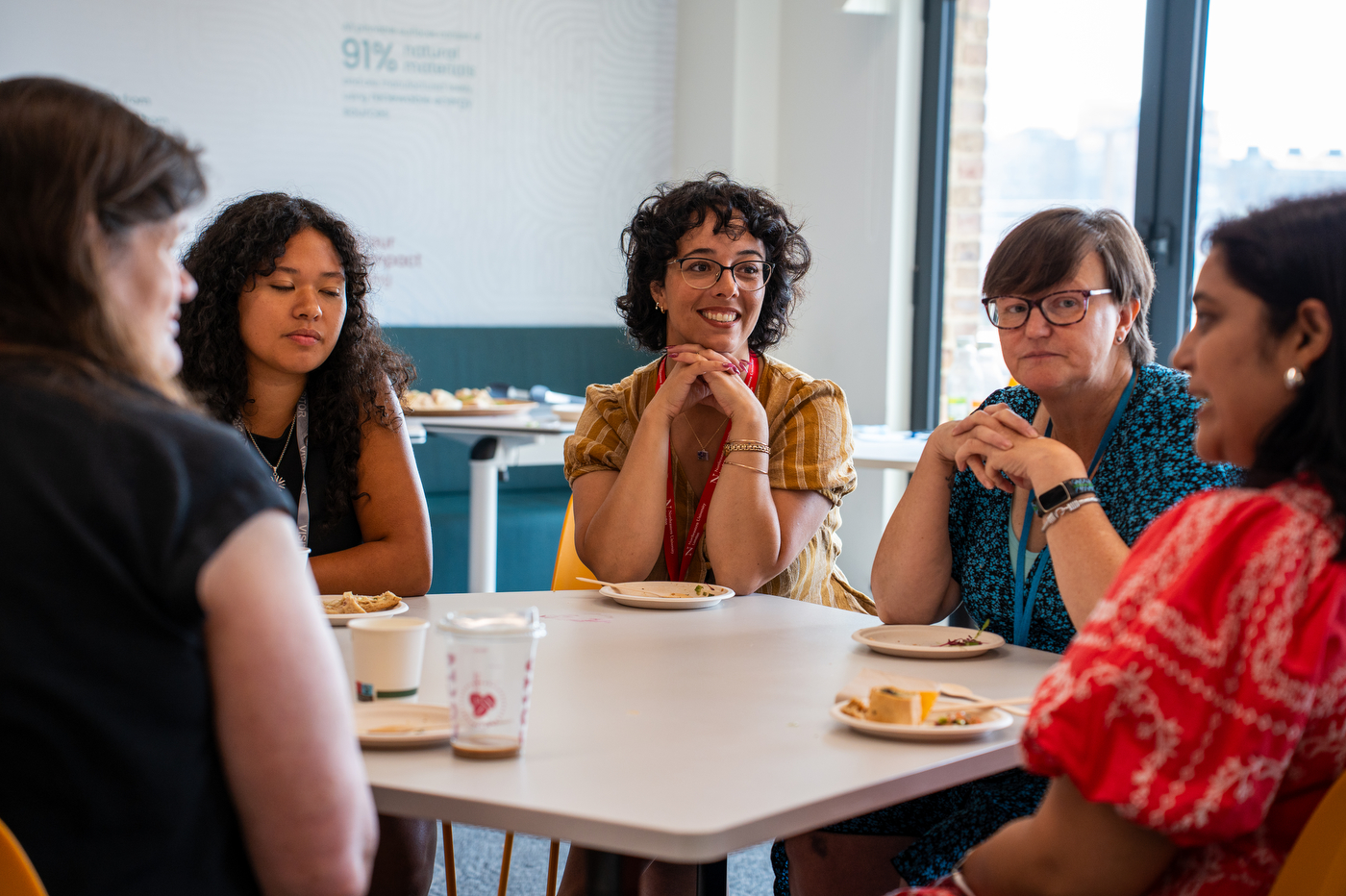 A small group sits around a table in conversation, coffee cups and plates in front of them.