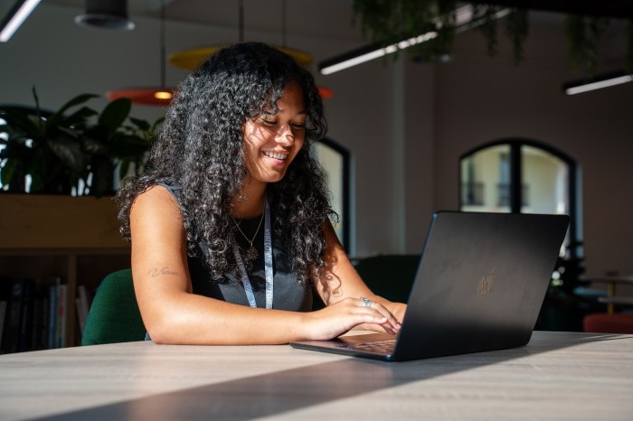 Patrina Danardatu smiles while typing on a laptop at a communal table in a bright, plant-filled workspace.