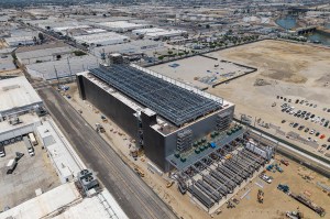 Aerial view of a large data center under construction, with cooling equipment and generators visible along the perimeter and undeveloped land surrounding the site.