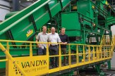 Three men stand on an elevated yellow walkway inside a Van Dyk Recycling Solutions facility, with green sorting and conveyor machinery behind them.