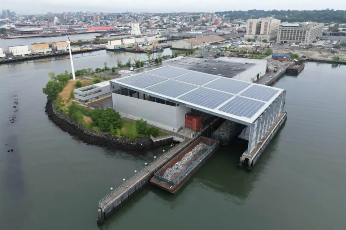 Aerial view of a waterfront recycling facility with a solar-paneled roof, a barge docked alongside it on the water, and a dense urban skyline in the background.