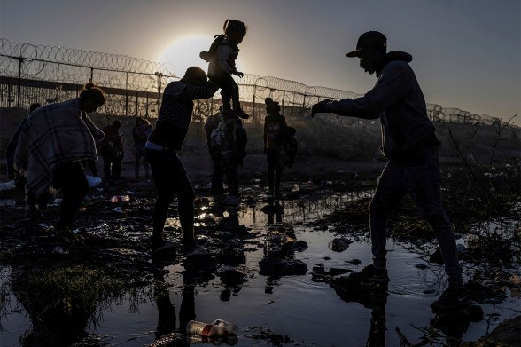 Migrants silhouetted against the sunrise as they cross a muddy riverbed near a razor-wire border fence.