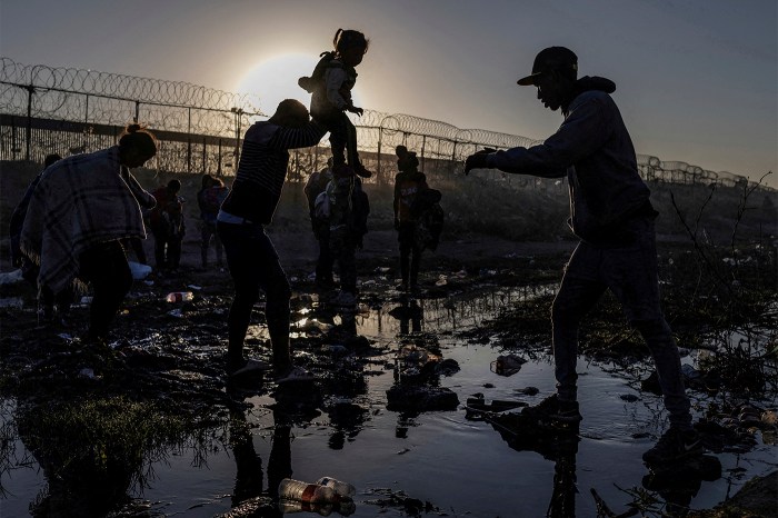 Migrants silhouetted against the sunrise as they cross a muddy riverbed near a razor-wire border fence.