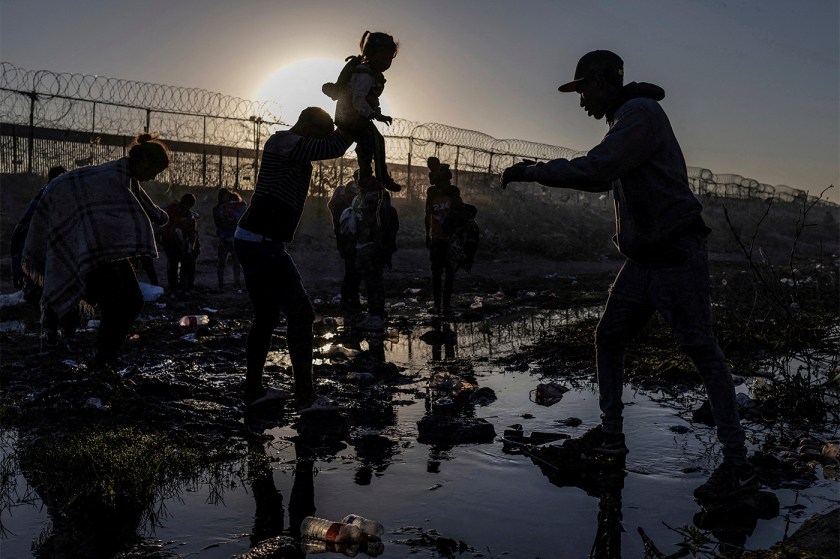 Migrants silhouetted against the sunrise as they cross a muddy riverbed near a razor-wire border fence.