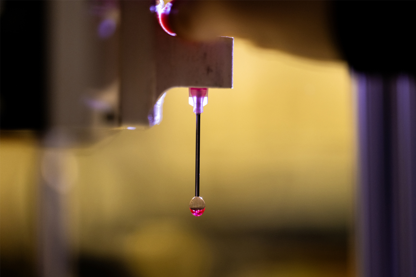 Close-up of a machine dispensing a small droplet of liquid, highlighted in pink light.