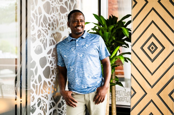 Yusuf Yassin, co-founder of Nuba and Nubian Markets, smiling in a blue patterned shirt in front of a painted wall and potted plant.