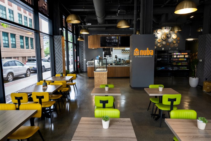 Wide interior view of Nuba by Nubian Markets, showing the service counter, Nuba logo sign, retail shelves, and gold pendant lighting, with floor-to-ceiling windows overlooking the street.