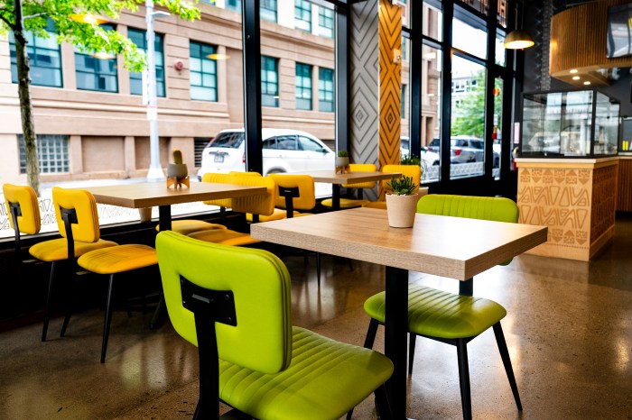 Green and yellow chairs at wooden tables inside Nuba by Nubian Markets, with street-facing windows in the background.