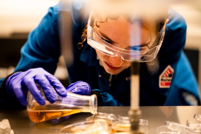 A student wearing protective gear and goggles carefully pours a liquid into another containter.