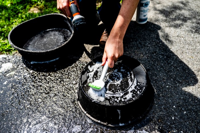 Close-up of a hand scrubbing a soapy feeding pan with a brush on a paved surface.