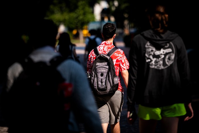 A crowd of people walk across campus, with one person seen from behind with a backpack and a bright red patterned shirt.