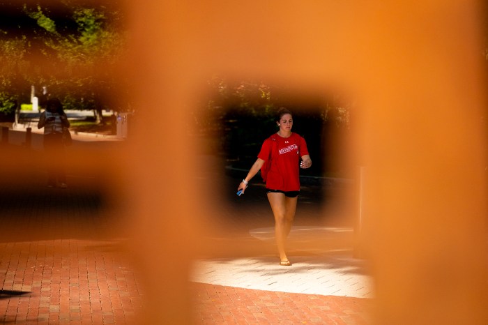 Person in a red shirt and black shorts walks along a sunlit brick path, framed through an out-of-focus orange object.

