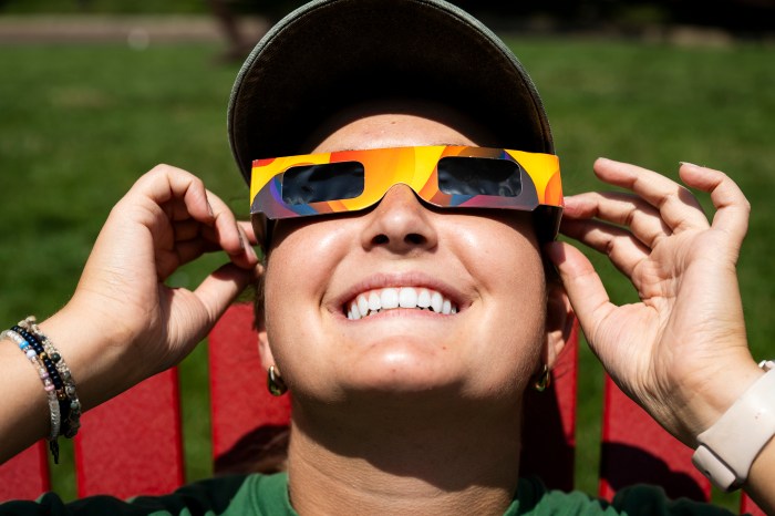 A student smiles while looking up through orange eclipse glasses outdoors.