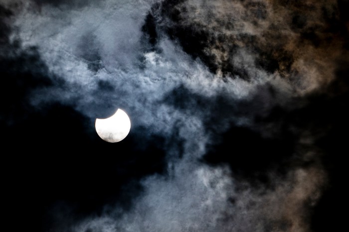 A partial solar eclipse visible through thick cloud cover, the moon's silhouette taking a bite out of the sun's disk.