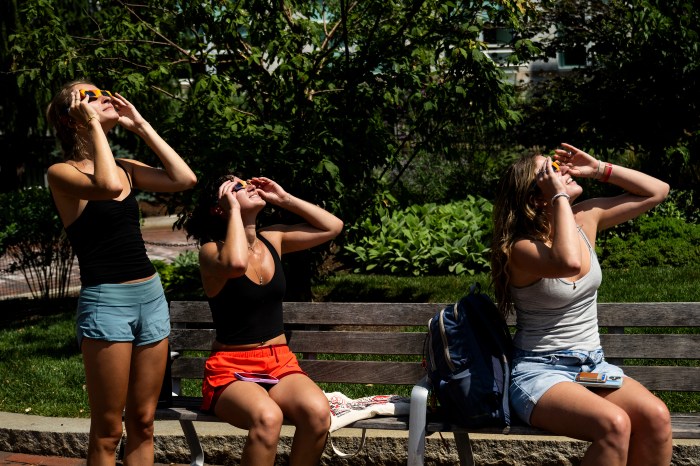 Three students view the partial solar eclipse through glasses while seated on a bench.