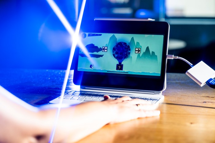 A hand rests on a laptop trackpad while a hot-air-balloon-themed puzzle game with directional arrows plays on screen, lit by a beam of blue light.