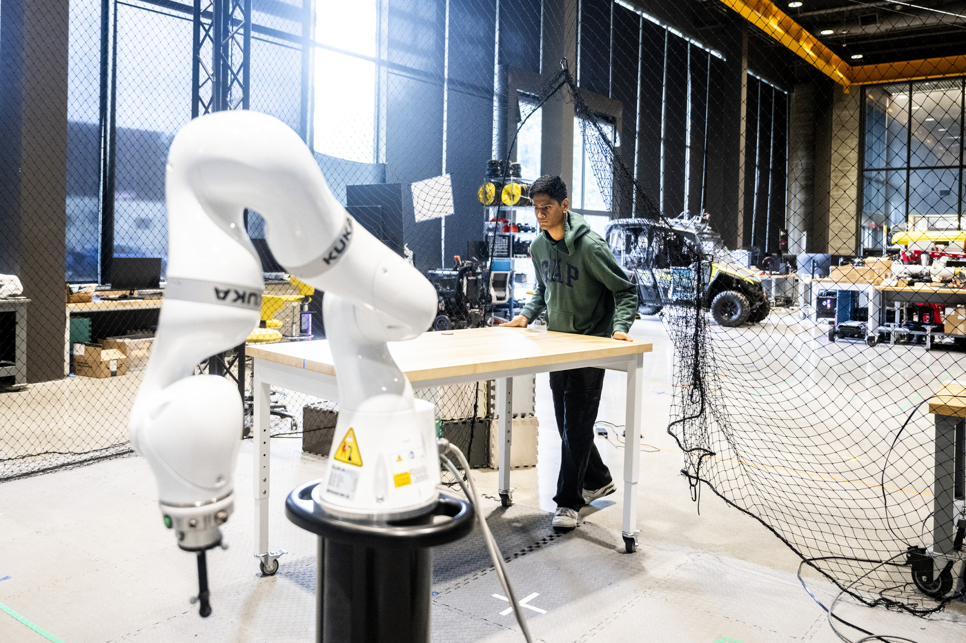 Haroon Hublikar works at a table near a white robotic arm in the EXP robotics high bay lab, with equipment and safety netting in the background.