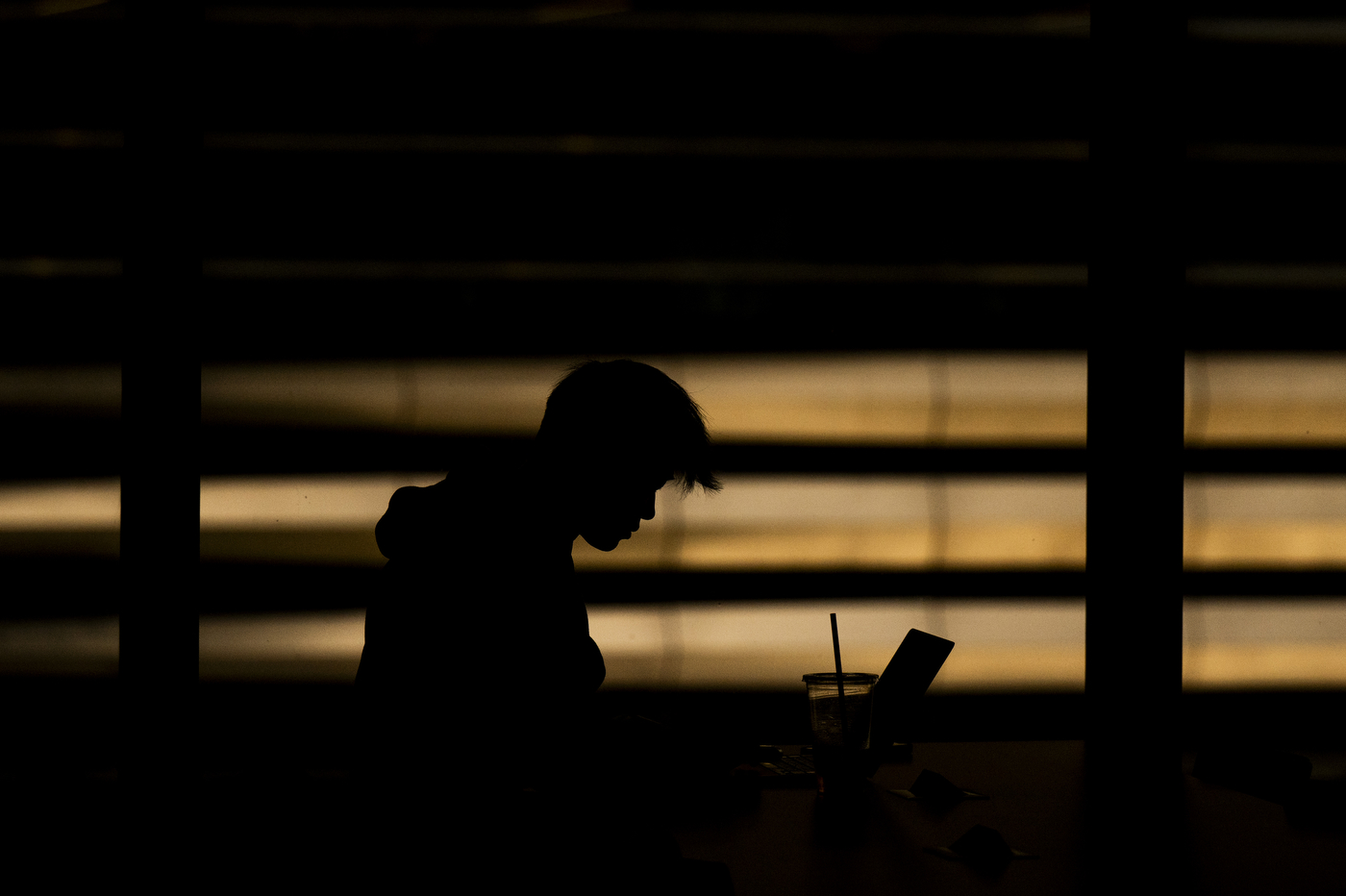 Silhouette of a student working on a laptop, backlit by sunlight through window blinds inside the ISEC building.