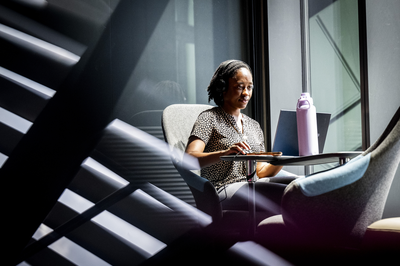 Woman wearing headphones works on a laptop at a small table beside a large window, with light streaming through window blinds in the foreground.
