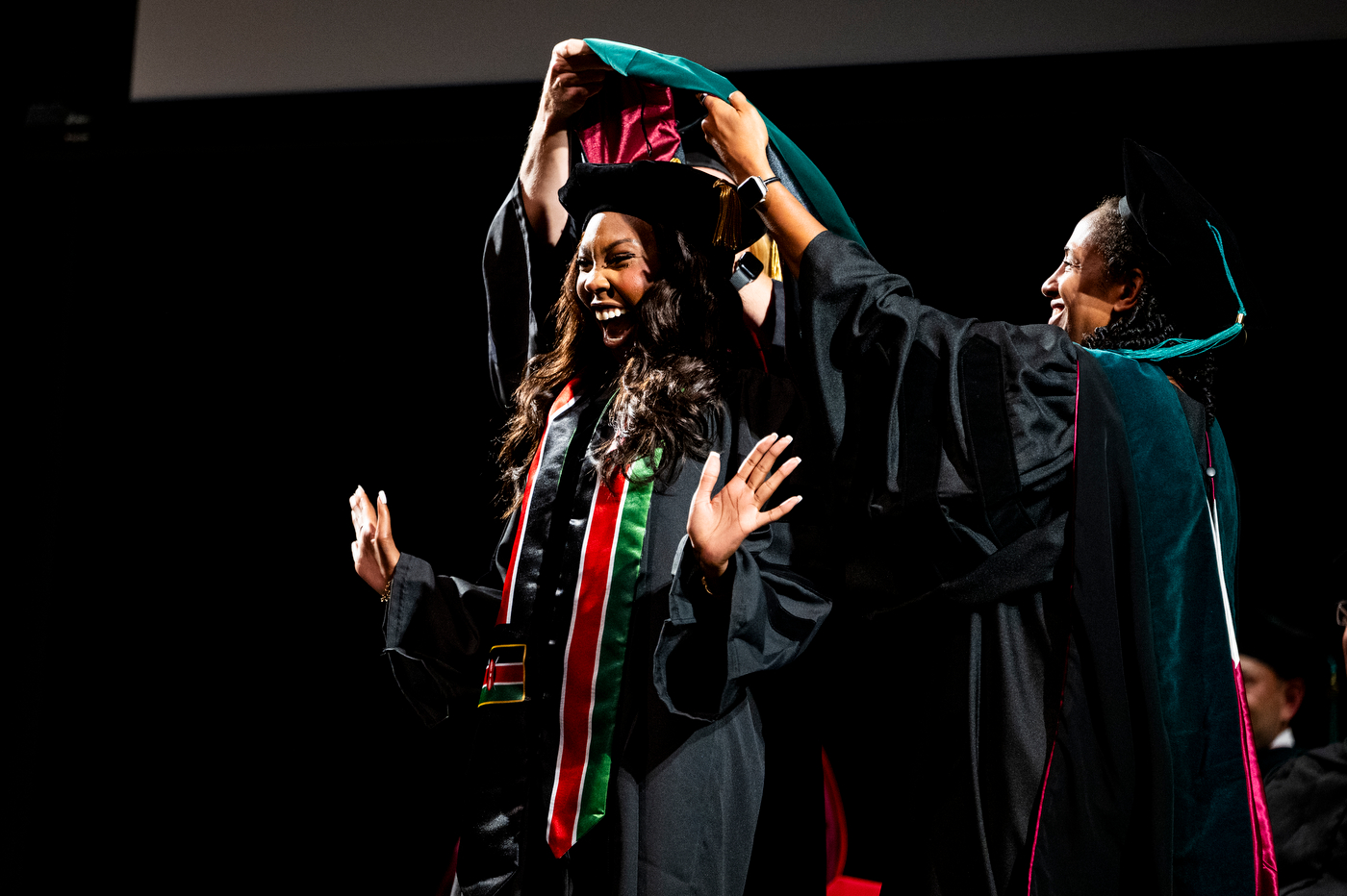 Smiling graduate raises her hands as a faculty member places a doctoral hood over her cap on stage, wearing a red and green ceremonial stole.