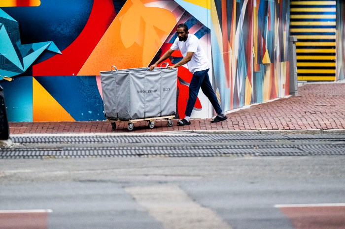 A student pushes a gray move-in cart down a brick walkway past a colorful abstract mural on campus.
