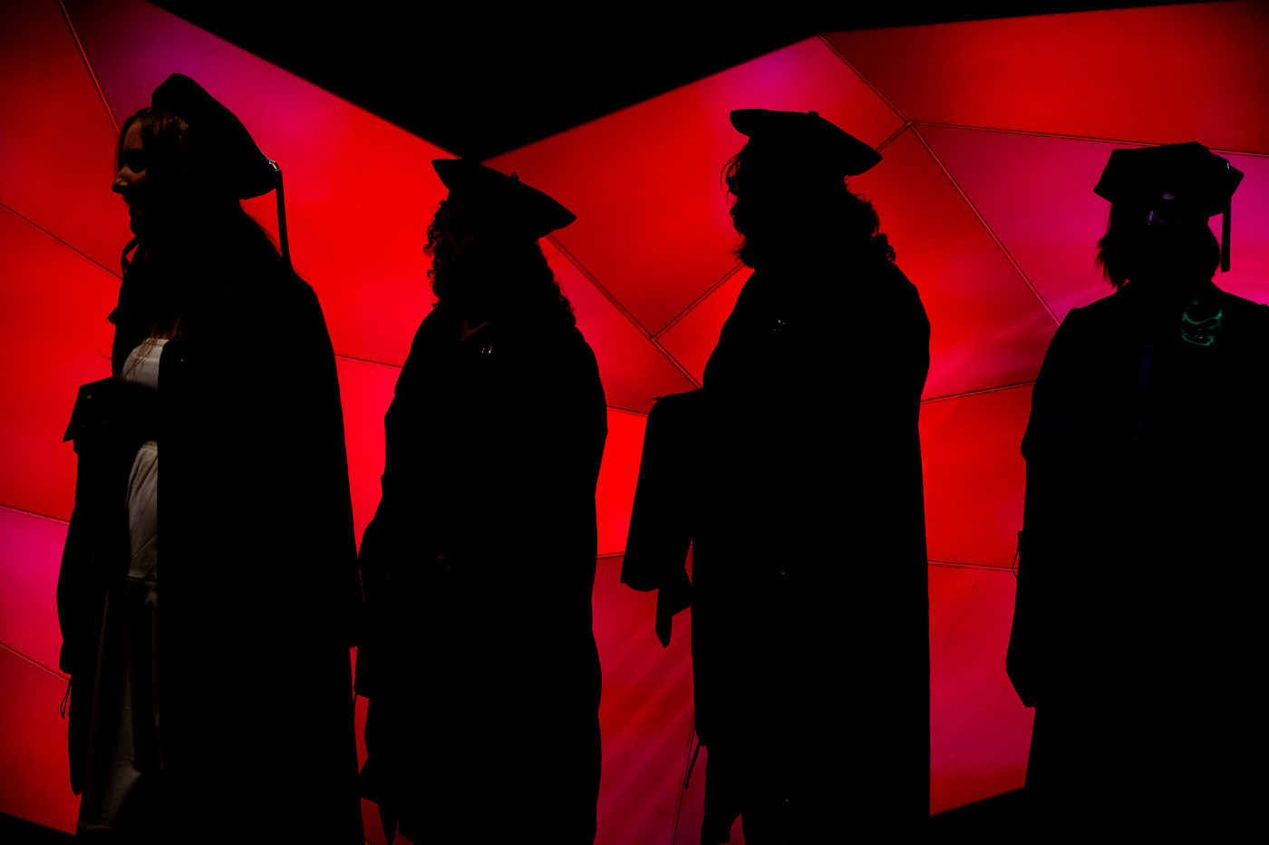 Silhouettes of graduates in doctoral regalia against a red-lit backdrop at the College of Professional Studies Doctoral Commencement Ceremony.