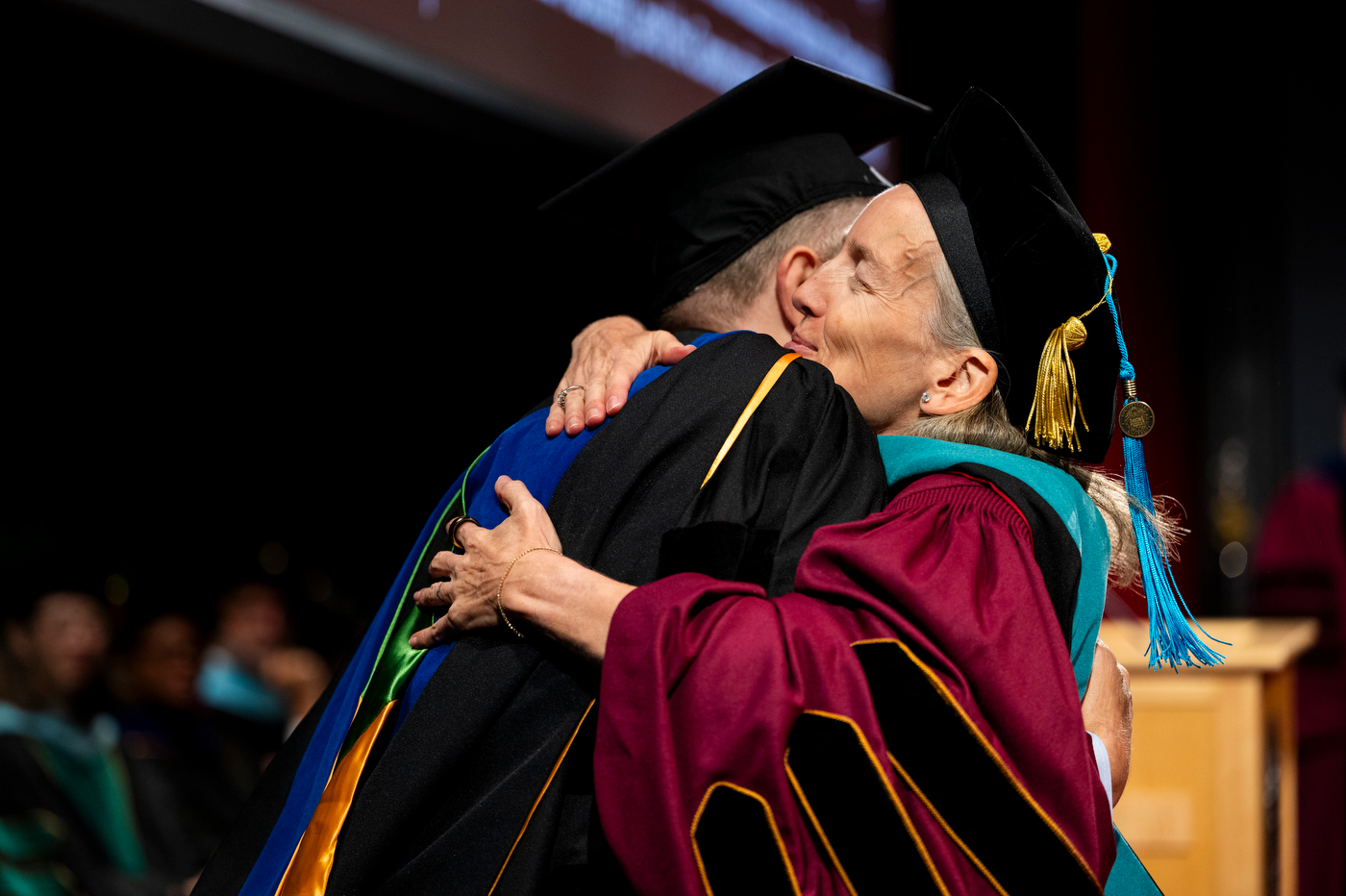 Two people hug on stage while wearing graduation regalia.