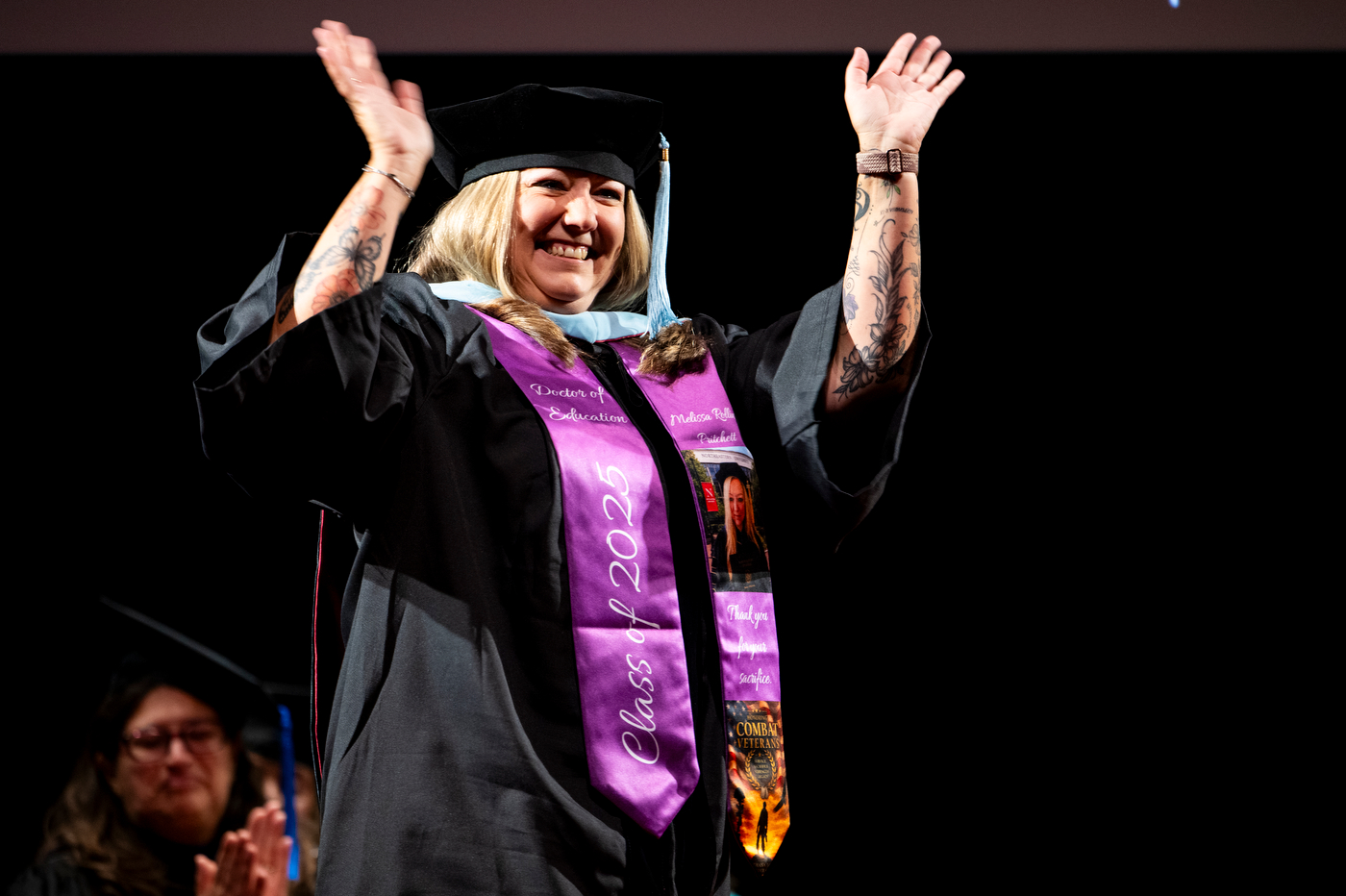 Doctoral graduate raises both arms and waves while crossing the stage in a purple sash during the College of Professional Studies Commencement.