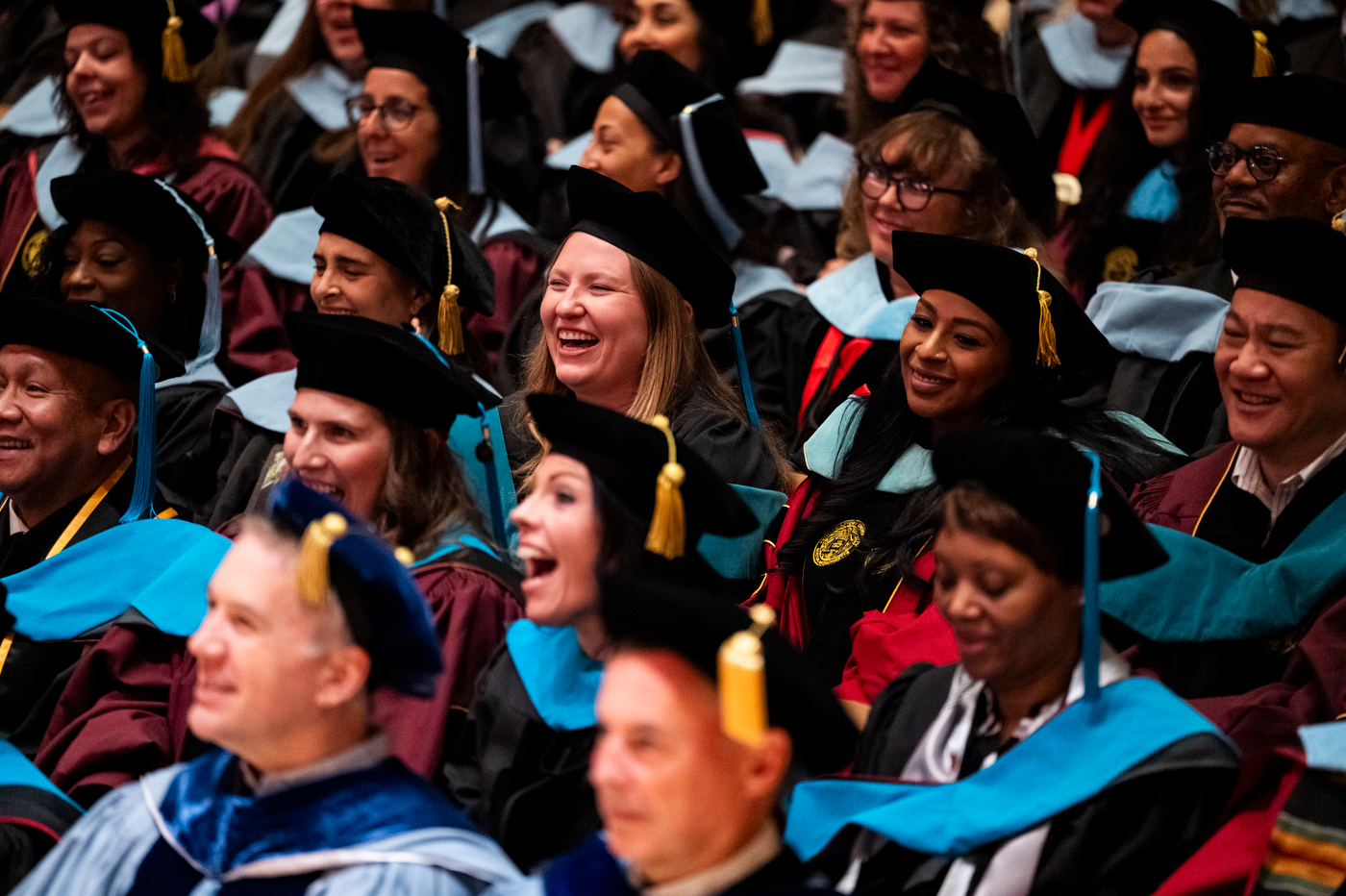 Several graduations in cap and gowns smile while seated in the audience.