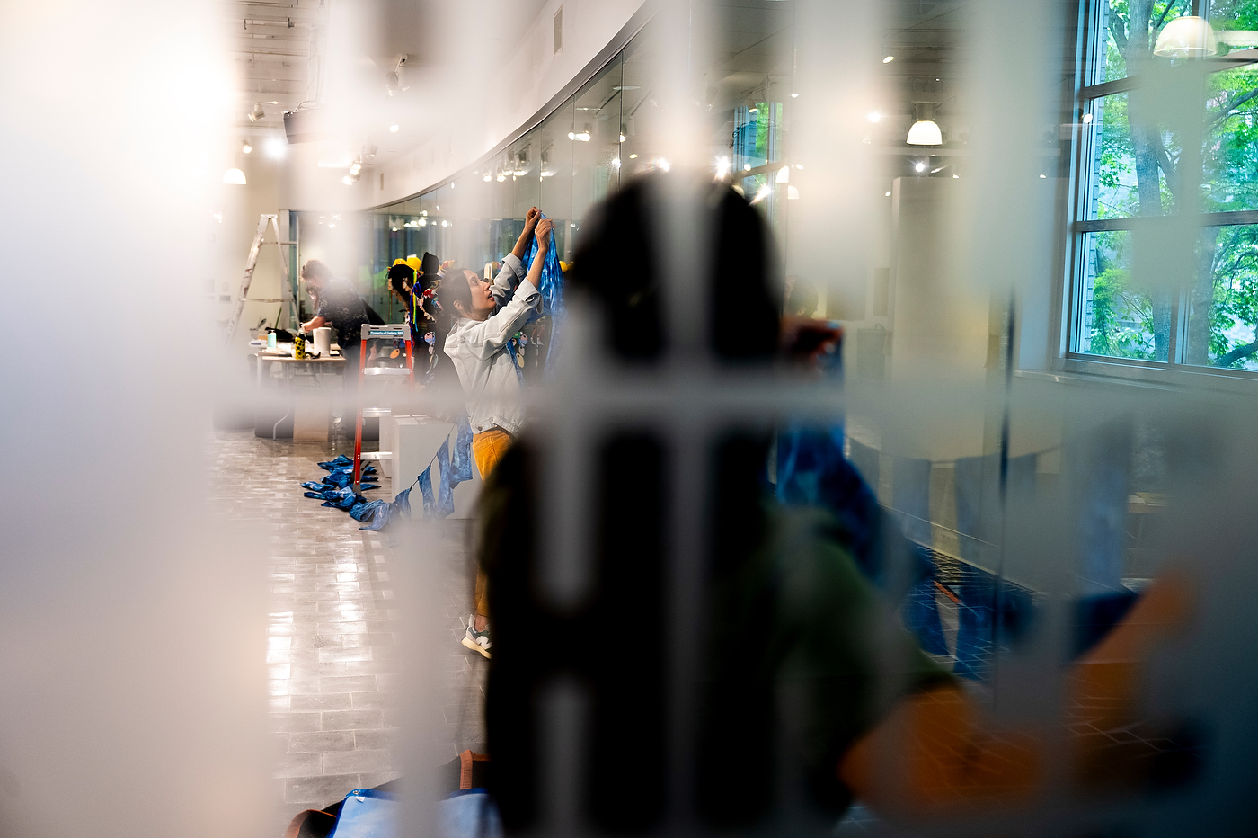 Blurred view through a reflective gallery window of staff and artists hanging framed artworks inside Gallery360.