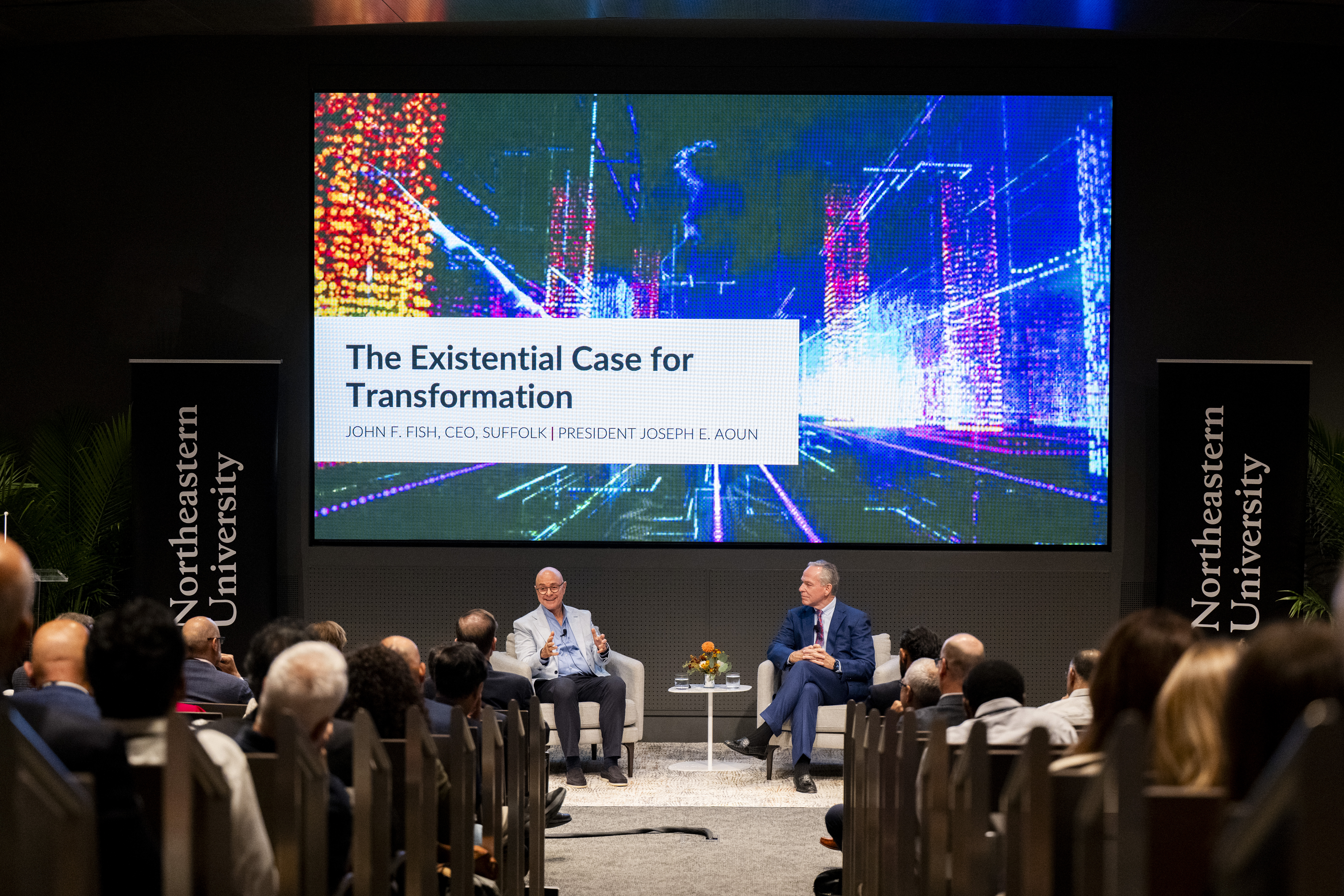 Wide view of President Joseph E. Aoun and John F. Fish sitting in armchairs at the fireside chat, with a title slide behind them that says 'Existential Case for transformation' and in front of them, audiences seated on either side of a center walkway.P