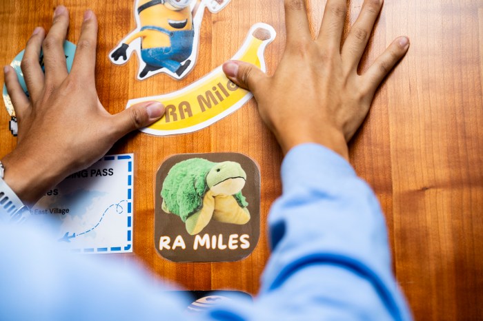 Close-up of hands placing "RA Miles" stickers, including a cartoon character and a plush frog magnet, on a wooden dormitory door.