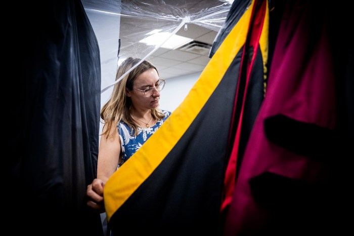 Chelsea Kryspin inspects a row of hanging academic gowns and gold-trimmed hoods while organizing faculty regalia for commencement.