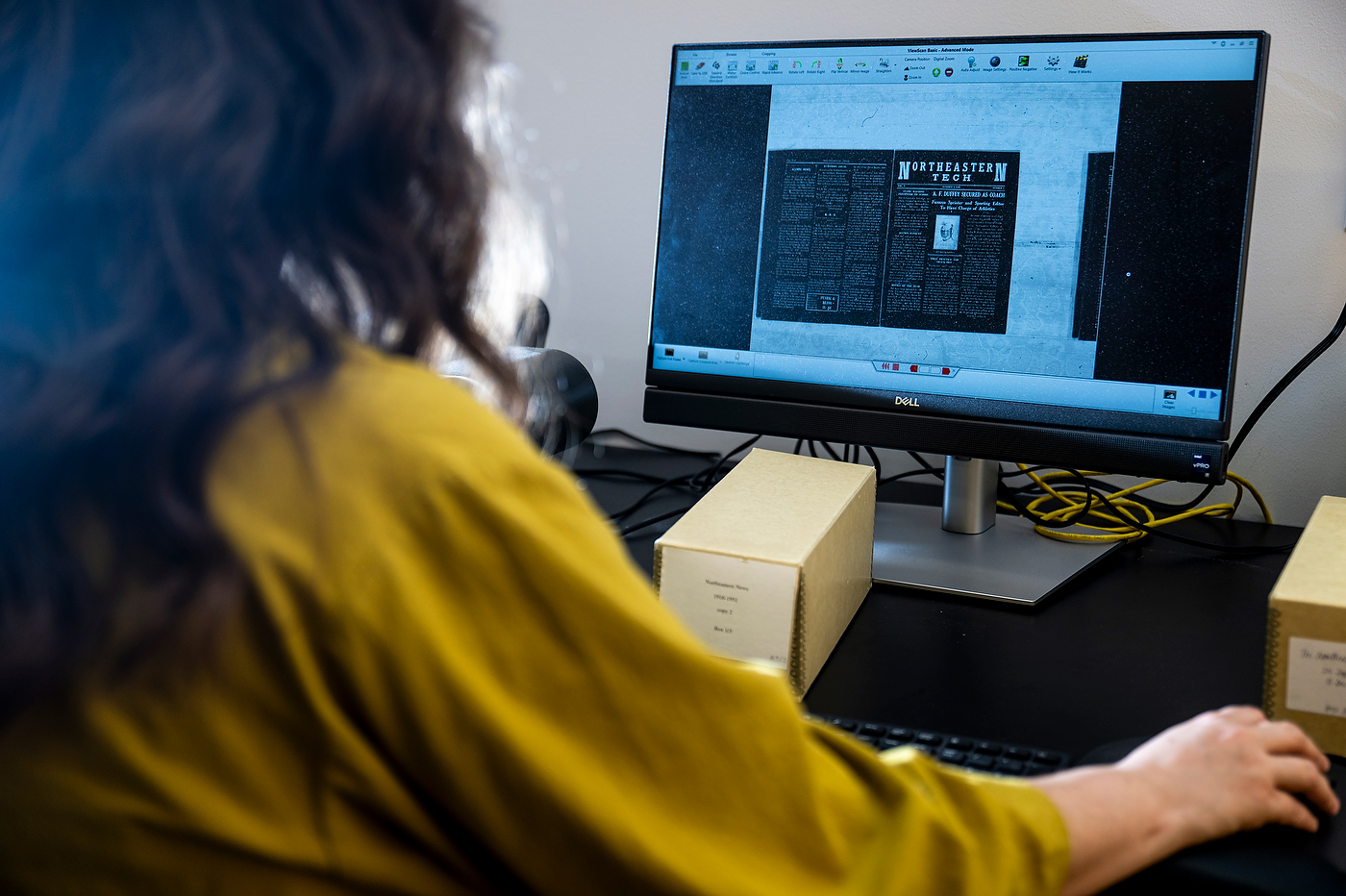 Molly Brown, seen from behind, viewing a scanned newspaper page on a monitor next to a labeled archive box.