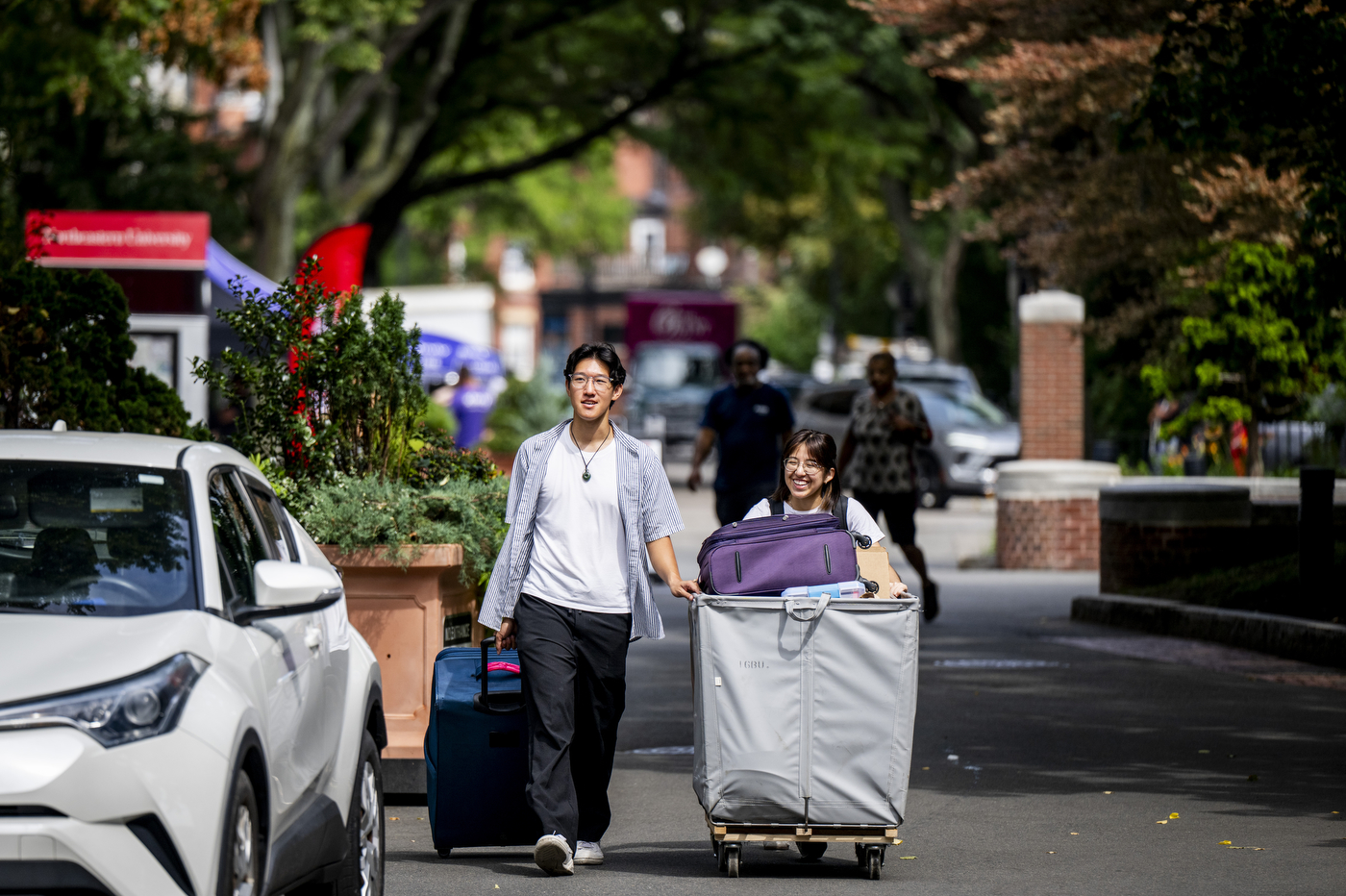 Michael Song and Michelle Montenegro push a wheeled cat full of their belongings down a tree-lined street.