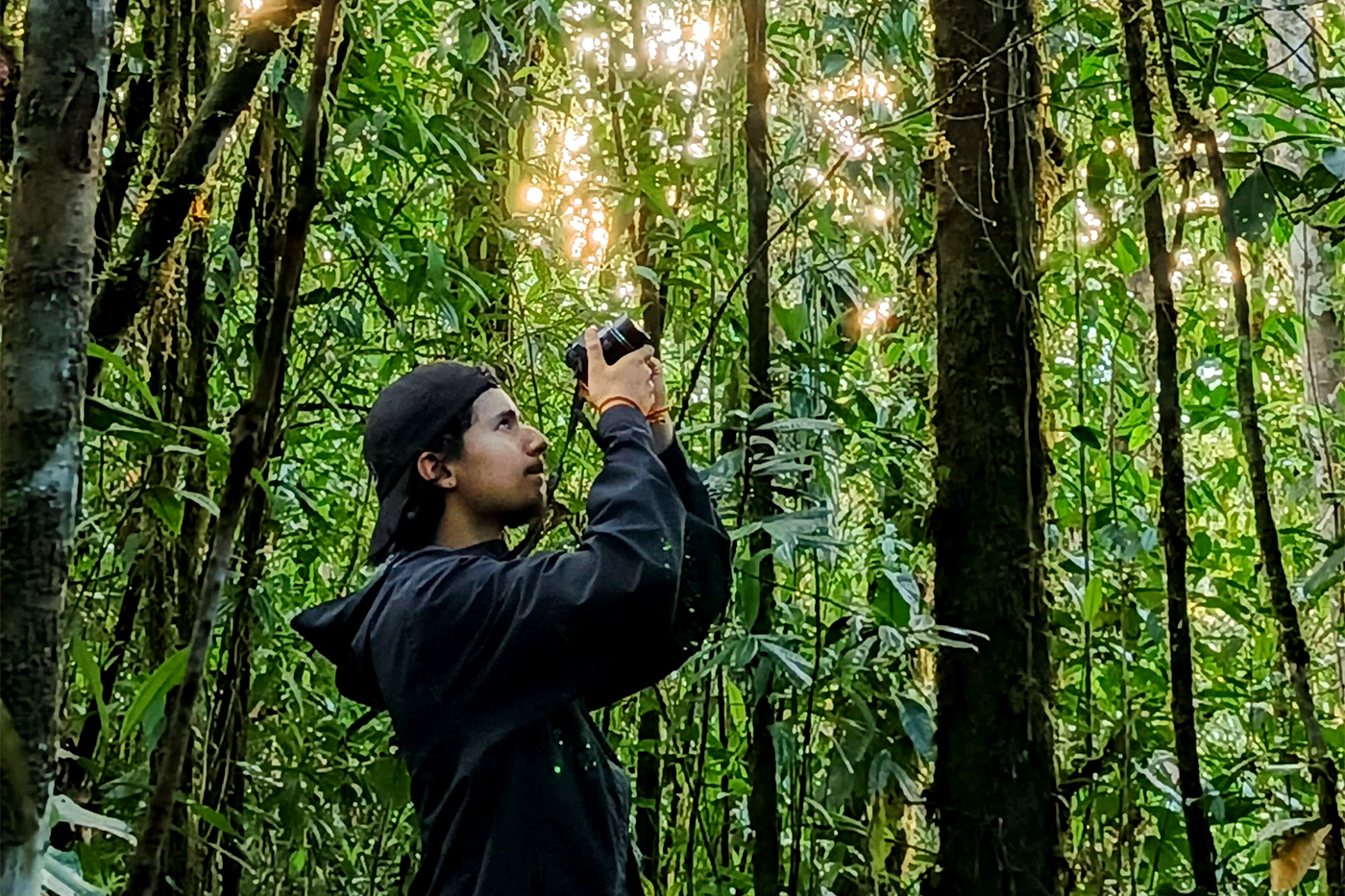 Adrian Baez holds a camera overhead, framed by shafts of sunlight breaking through the canopy.