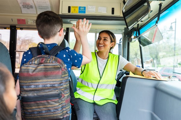 A school bus driver wearing a neon vest giving a high five to a young student.