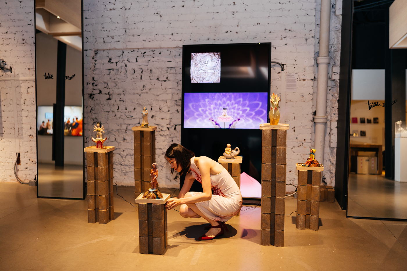 A person in a pink dress crouches to arrange a small figurine atop a stone pedestal, surrounded by similar sculptures displayed on pillars in front of a brick wall with video screens.